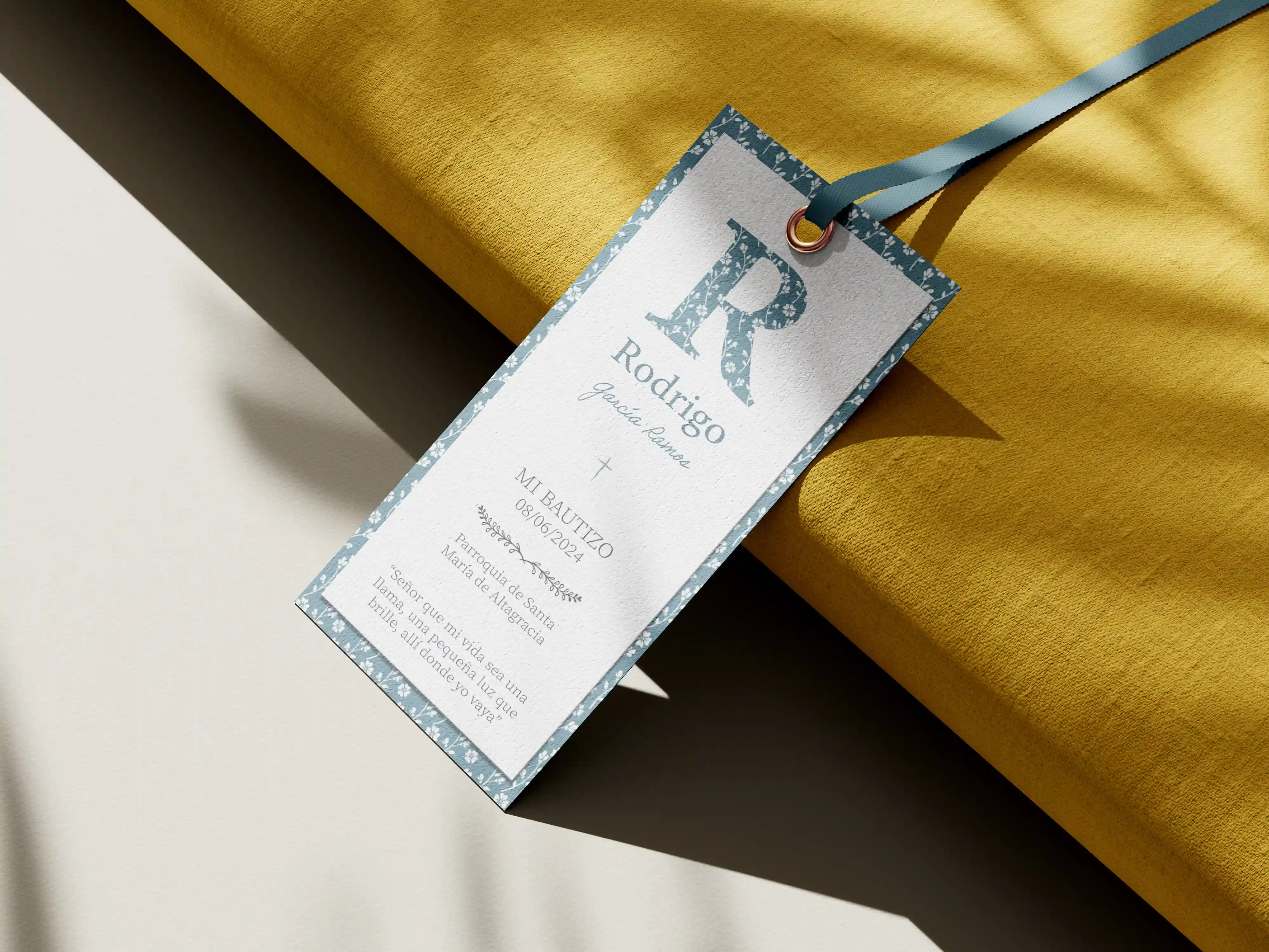 Recordatorio de bautizo personalizado con marco floral azul y cinta, colocado sobre una tela dorada.